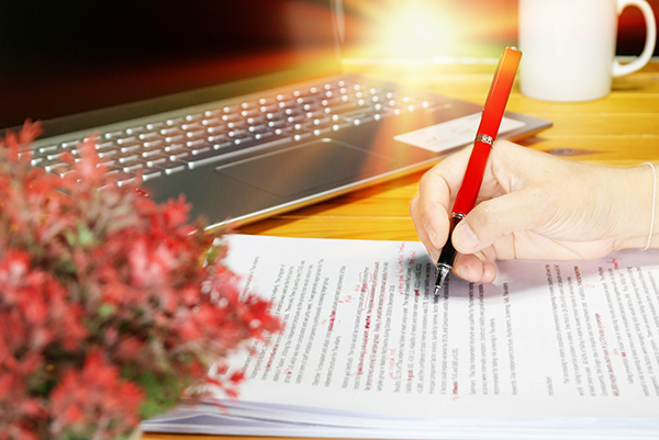 Decorative image showing a hand manually proofreading a printed page in front of a laptop